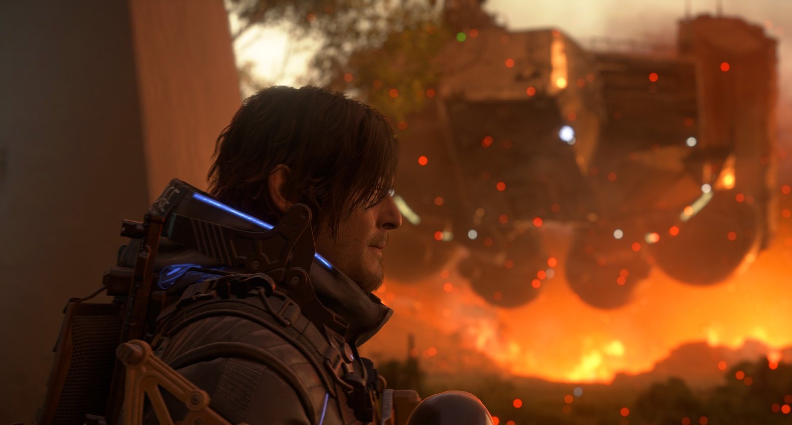 A profile shot of Sam Porter Bridges from the video game Death Stranding 2. He is wearing his Bridges suit and looking towards a fiery, chaotic landscape in the distance, lit by orange and red light.